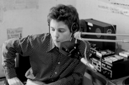 Blurred black and white photo of young man wearing polka dot shirt at radio mixing desk with microphone