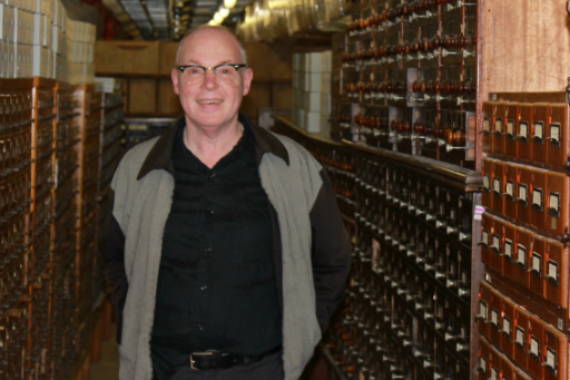 Portrait of man wearing glasses, grey vest and black shirt standing in a basement in front of wooden catalogue drawers