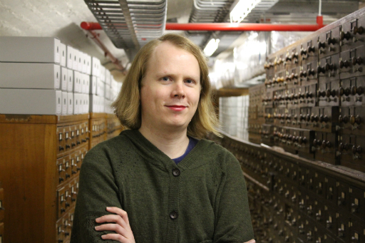 Portrait of person wearing red lipstick and brown jacket standing in a basement in front of wooden catalogue drawers